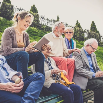 Foto zeigt eine Gruppe von Menschen im Freien auf einer Bank sitzend. Die Frauen und Männer sind fortgeschrittenen Alters. Sie unterhalten sich oder nutzen ihre digitalen Endgeräte.